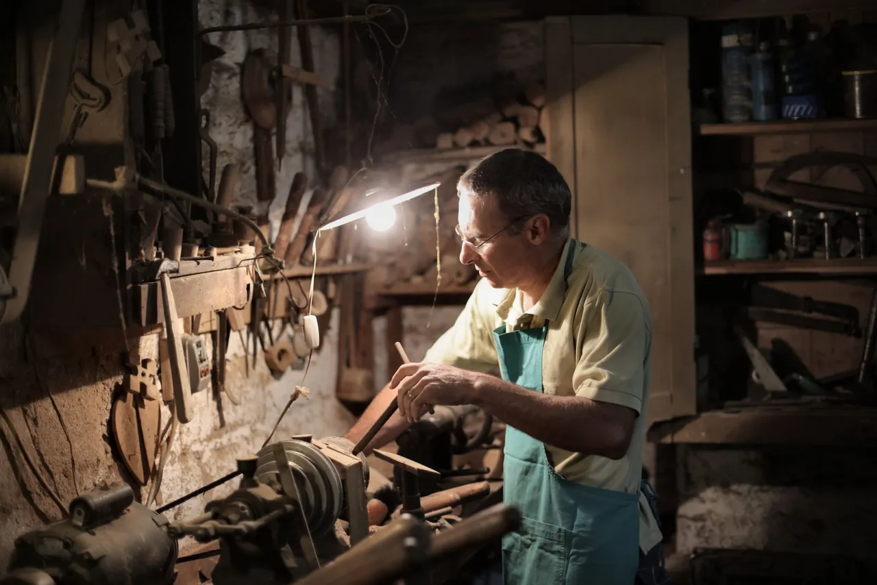 Handwerker in einer kleinen, schwach beleuchteten Werkstatt arbeitet konzentriert an einer Drehbank, an der Wand hängt Werkzeug