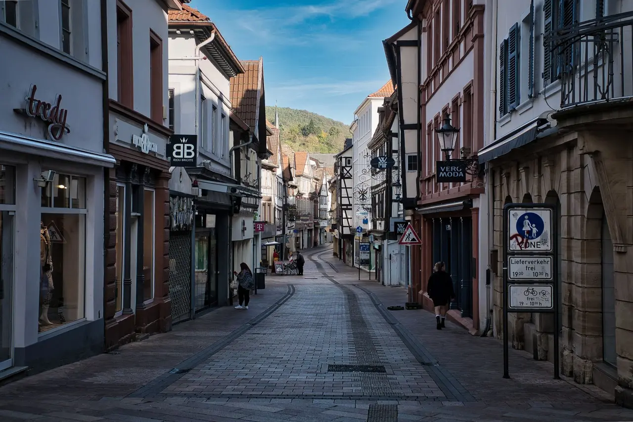Gasse einer europäischen Kleinstadt mit Geschäften auf beiden Seiten, im Hintergrund bewaldete Hügel