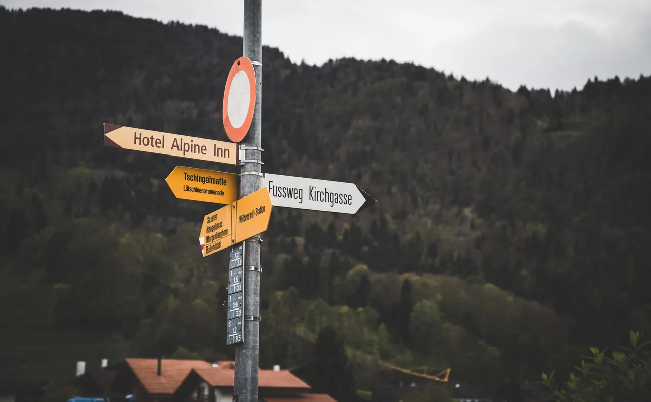 Wegweiser mit mehreren gelben Richtungsschildern vor bewaldeten Hügeln, Sinnbild für lokale Reichweite im Bezirk
