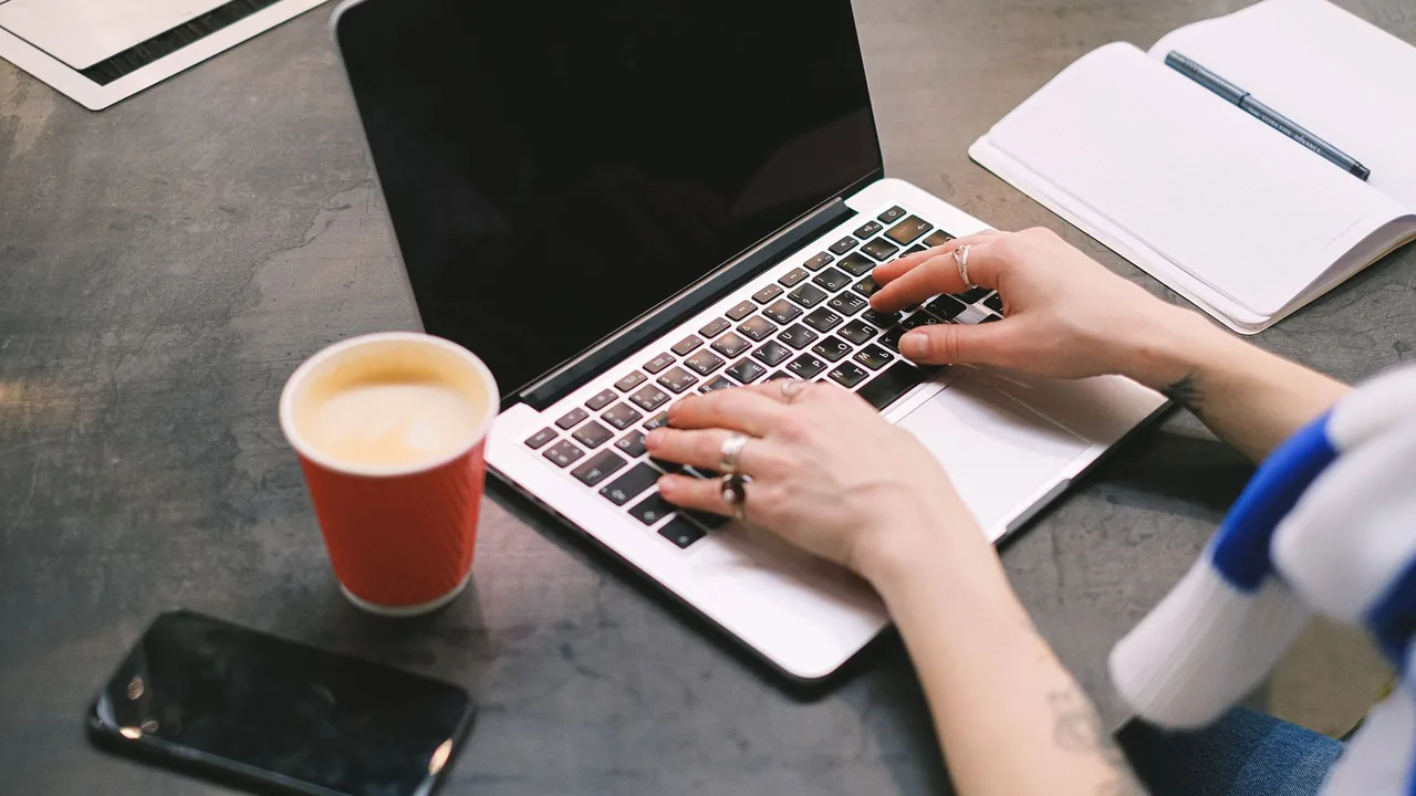 Hände tippen an einem Laptop an einem aufgeräumten Schreibtisch mit Kaffee und Notizbuch als Sinnbild für die Website-Erstellung an einem Tag