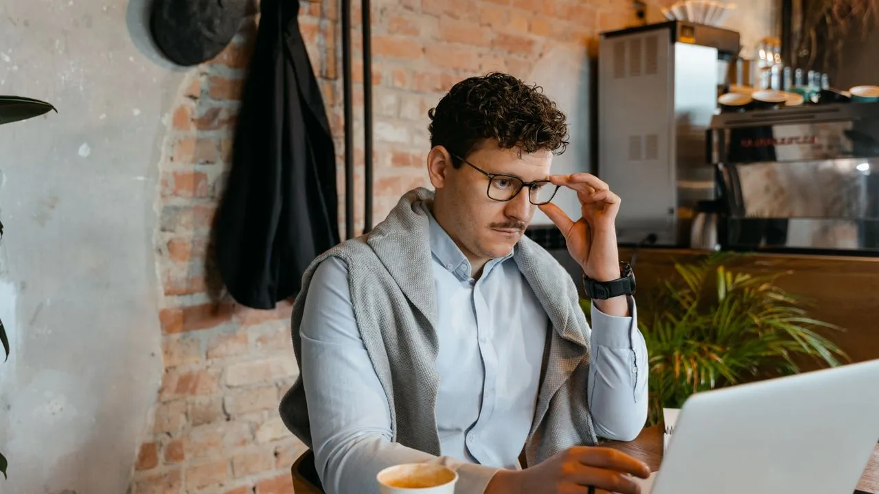 Kleinunternehmer sitzt nachdenklich mit Brille vor dem Laptop und überlegt, ob sich eine neue Website rechnet