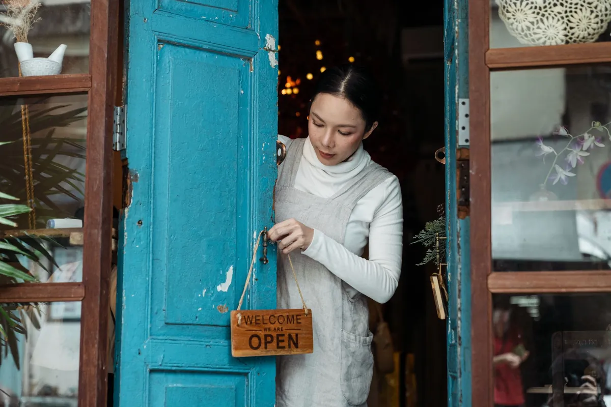 Frau hängt an einer blau gestrichenen Holztür ein Holzschild mit der Aufschrift „Welcome, we are open“ auf, Sinnbild für den Livegang einer neuen Website
