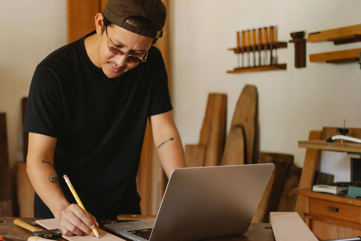Tischler in seiner Werkstatt notiert mit einem Bleistift Anmerkungen auf einem Zettel, vor ihm ein aufgeklappter Laptop, Sinnbild für die Rückmeldung zum Website-Entwurf