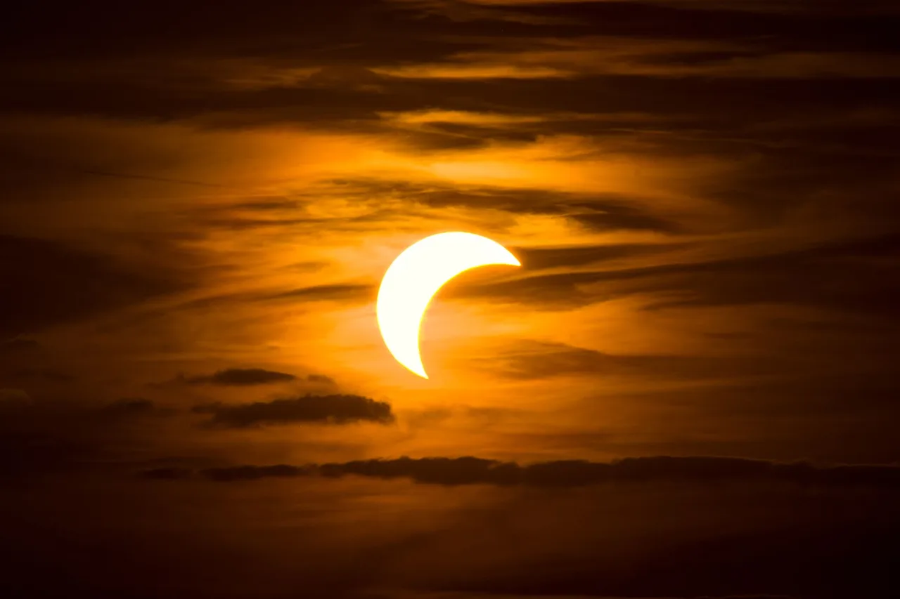 Partielle Sonnenfinsternis als leuchtende Sichel hinter dramatischen Wolken am orangefarbenen Abendhimmel