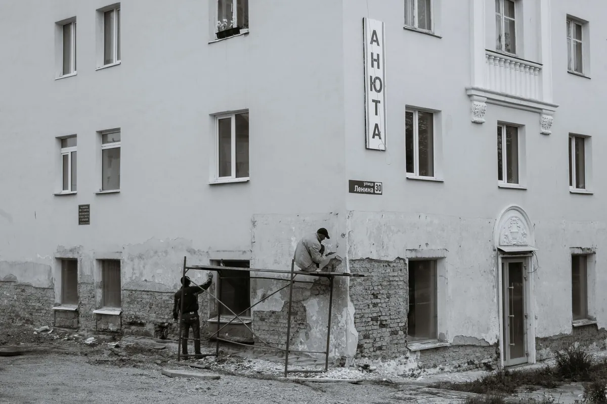 Schwarzweißfoto zweier Arbeiter auf einem Gerüst, die den bröckelnden Putz an einer alten Hausfassade ausbessern: Flicken hält, solange die Mauer dahinter trägt